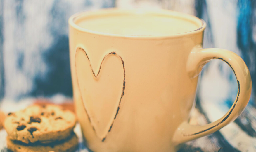 Close up of a peach coffee cup with a heart engraved on the front, sat next to two chocolate chip cookies.