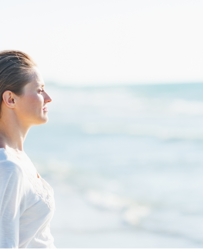 Woman in white tunic stands staring out to sea on a sunny day