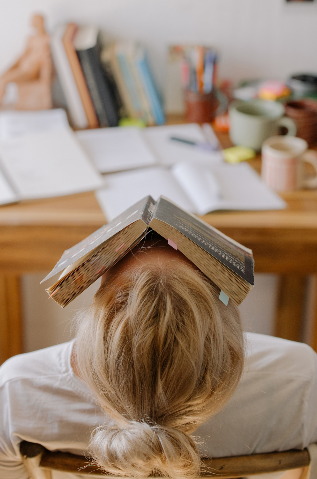 Woman lies back in chair with book covering her face in front of a cluttered desk.