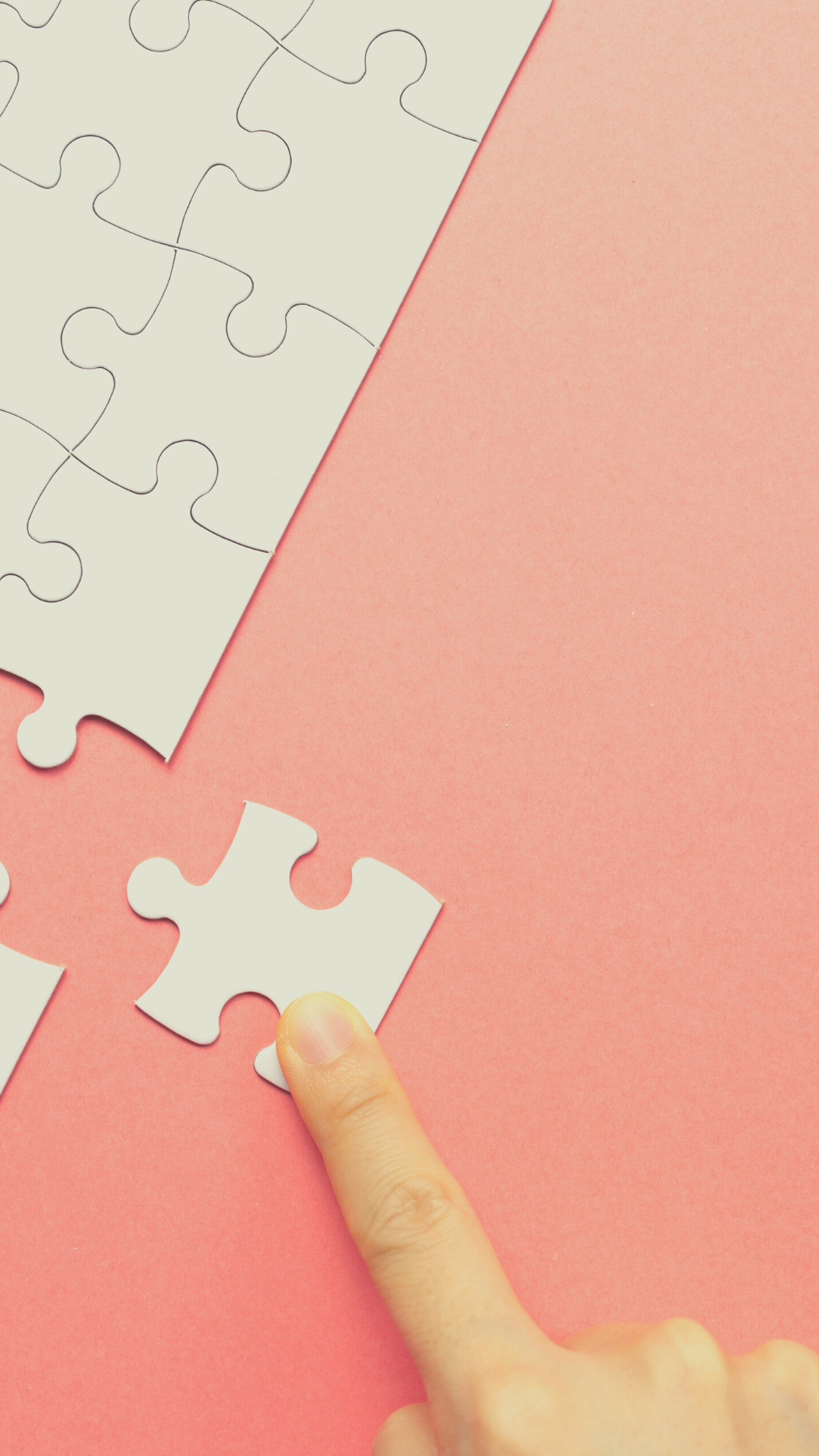 Close up of a hand placing a white jigsaw piece into a jigsaw against a pink background