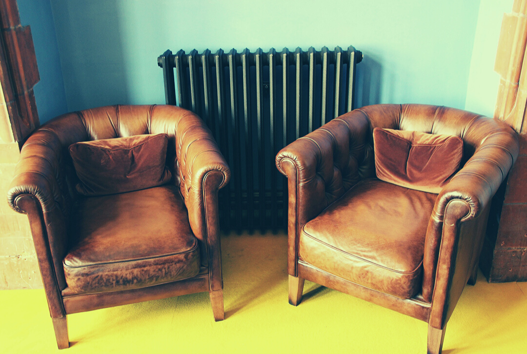 Two brown leather armchairs sit side by side a black retro radiator. On a yellow floor against a turquoise wall.