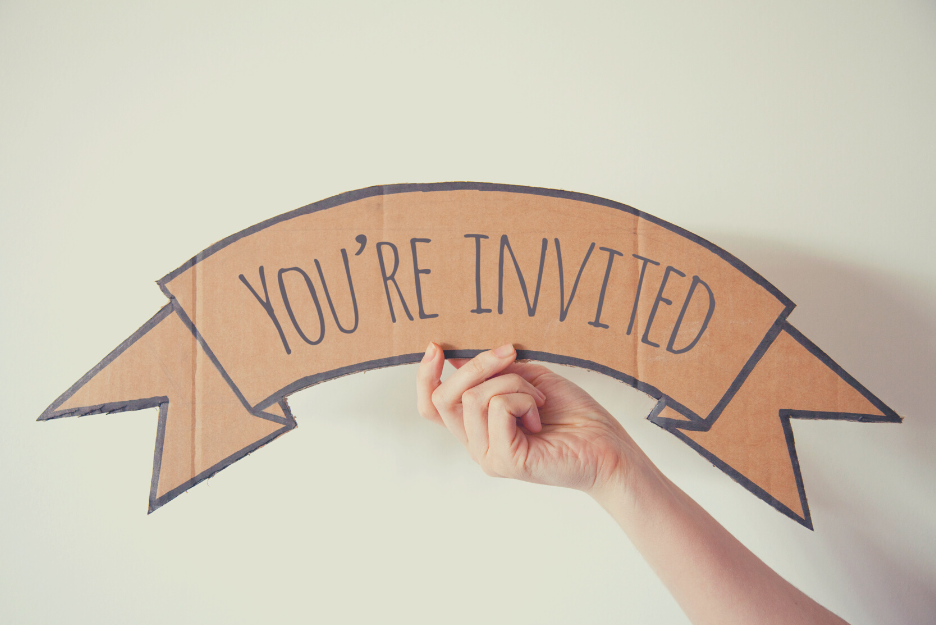 A hand holds up a cardboard banner that reads "YOU'RE INVITED'