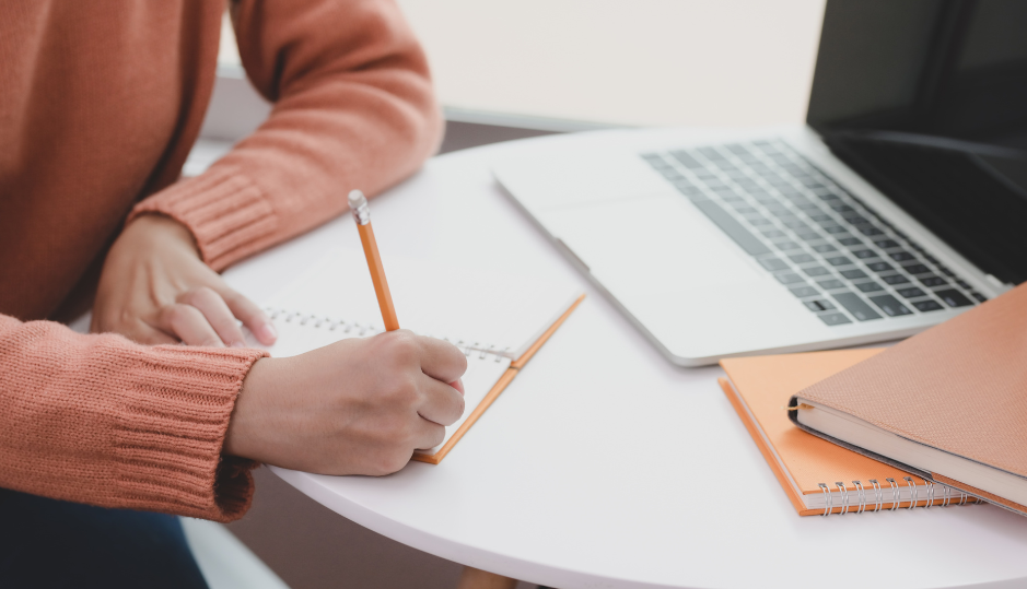 Woman in peach jumper writes in pencil on a notepad. She's sitting at a white round desk with her laptop and two larger notepads.