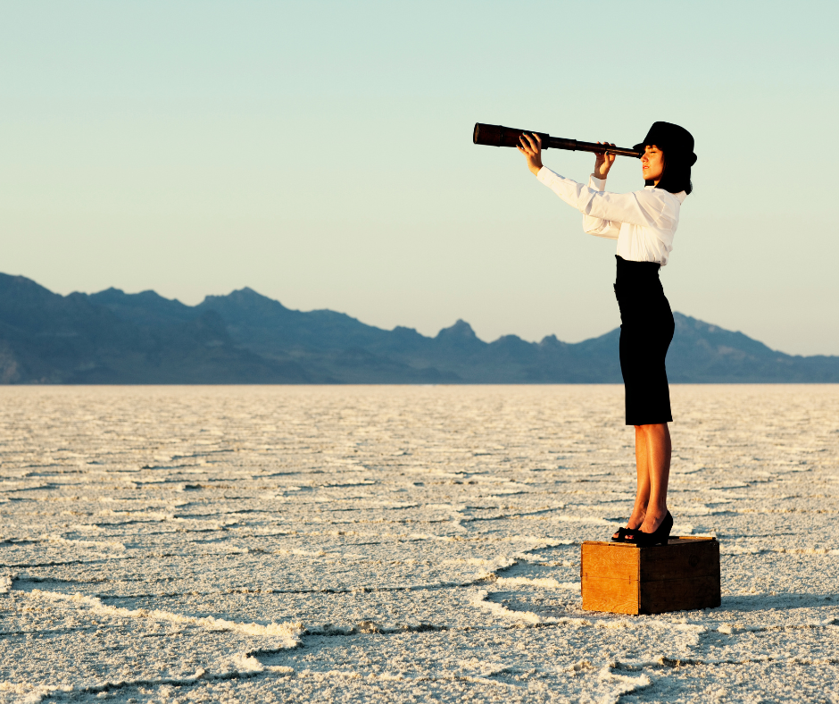 Woman in skirt suit stands on a box in the desert looking into the distance using a telescope. Symbolising the long term vision for her counselling business.