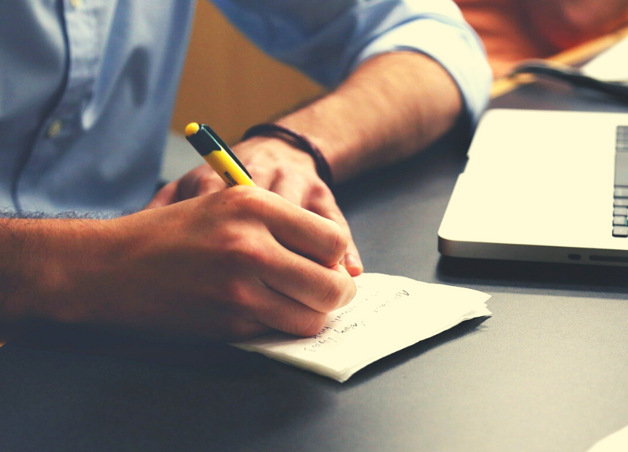 Man writing session notes with a yellow pen on black desk next to laptop