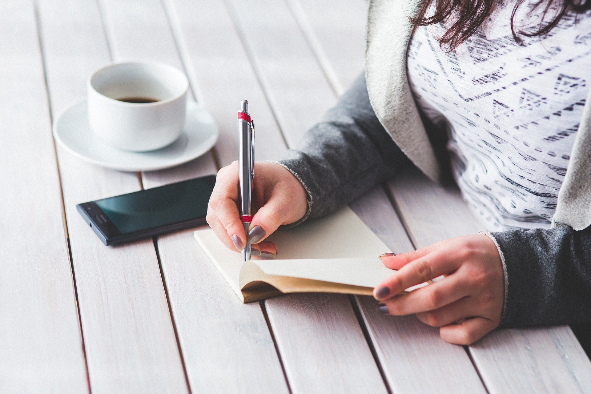 Woman writing notes in notebook. Coffee cup and mobile phone sit on white table.