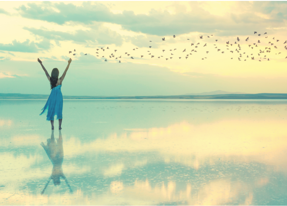 Woman in blue dress standing with arms up on a beach as a flock of birds fly away