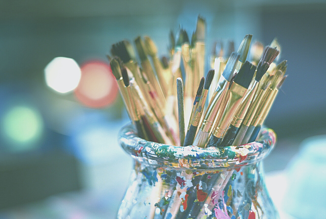 Paintbrushes in a paint covered jar against a blue background.