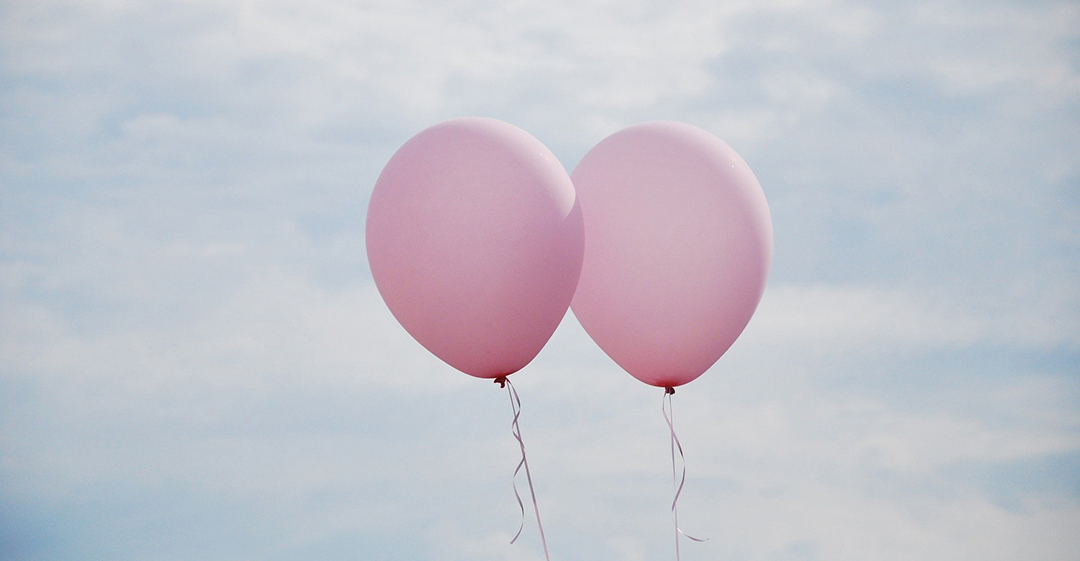 Two pink balloons floating together in a blue sky.