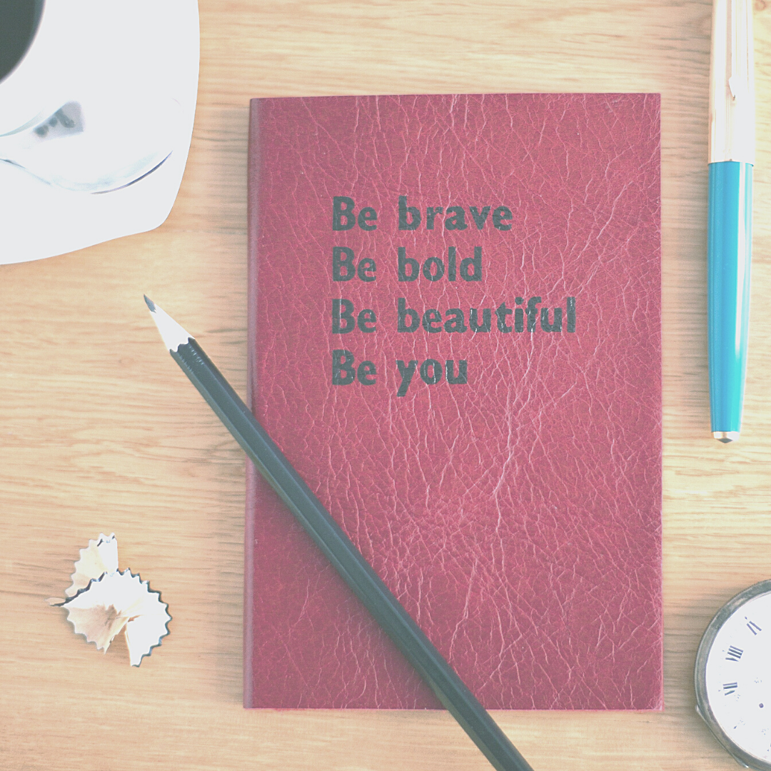 Red journal on a desk with blue pen, black pencil and pocket watch. Cover reads Be brave, Be bold. Be beautiful. Be you.